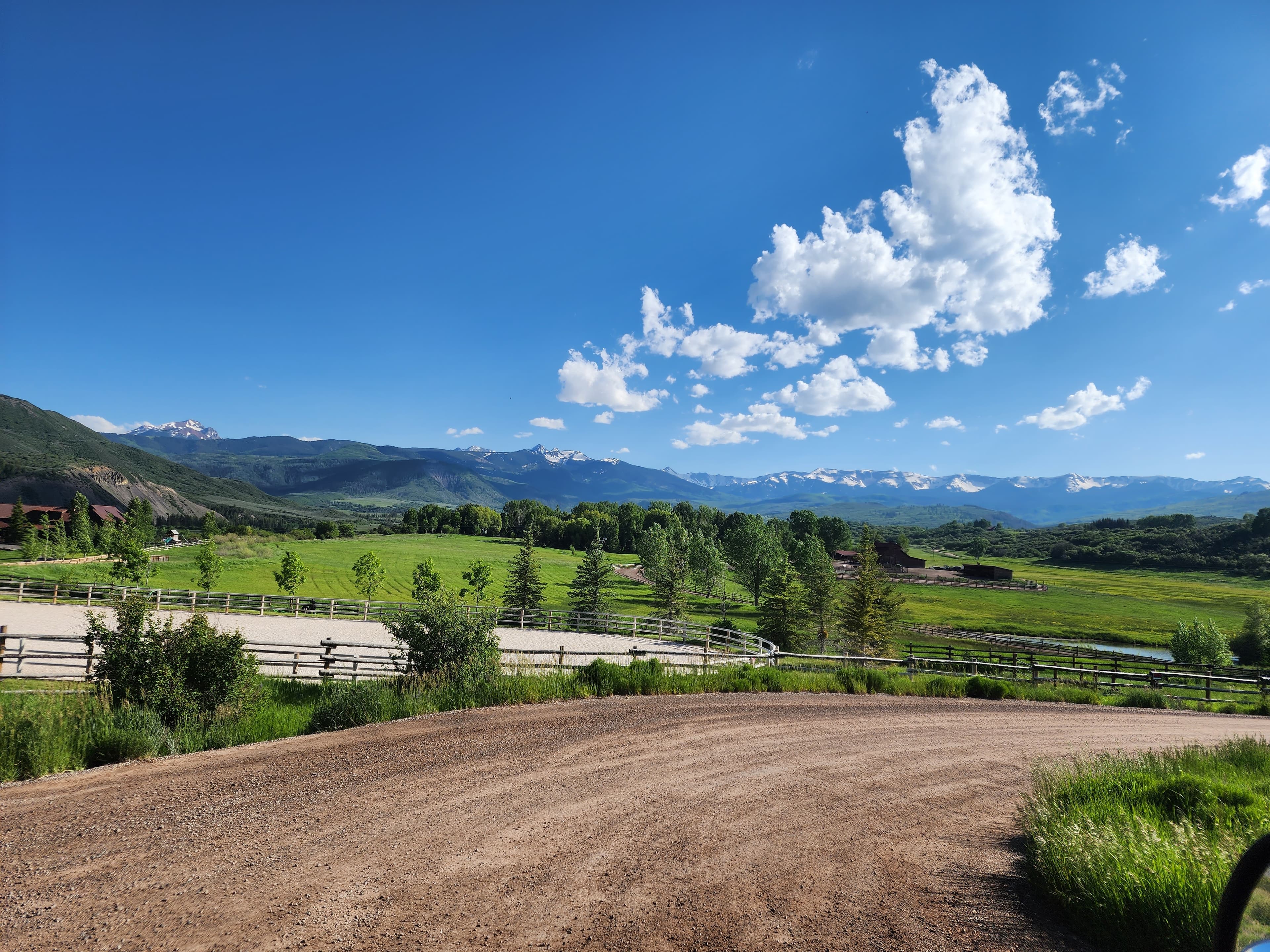 Scenic green valley with a horse arena and distant snow-capped mountains under blue skies.