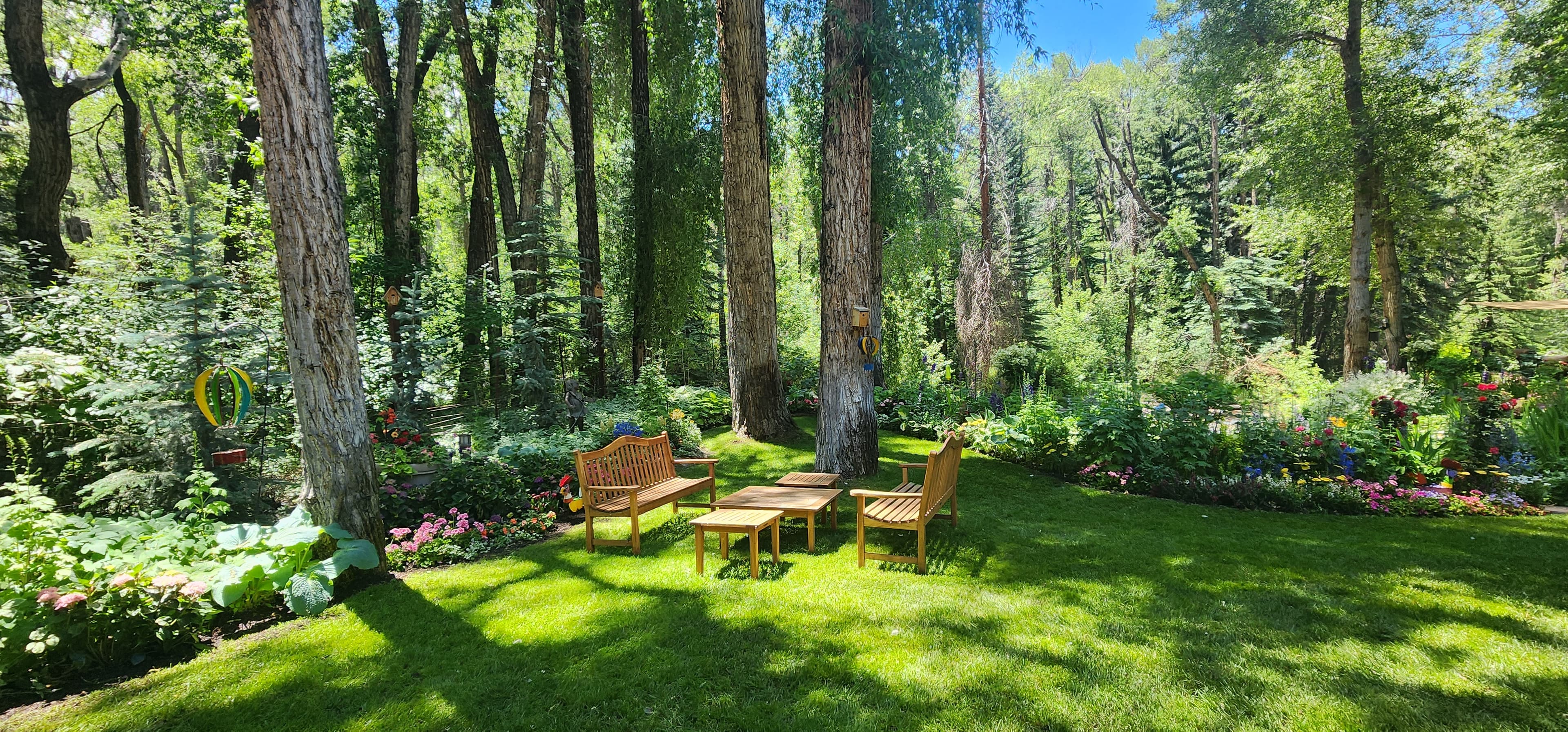 Wooden furniture on a sun-dappled lawn surrounded by tall trees and colorful garden flowers.