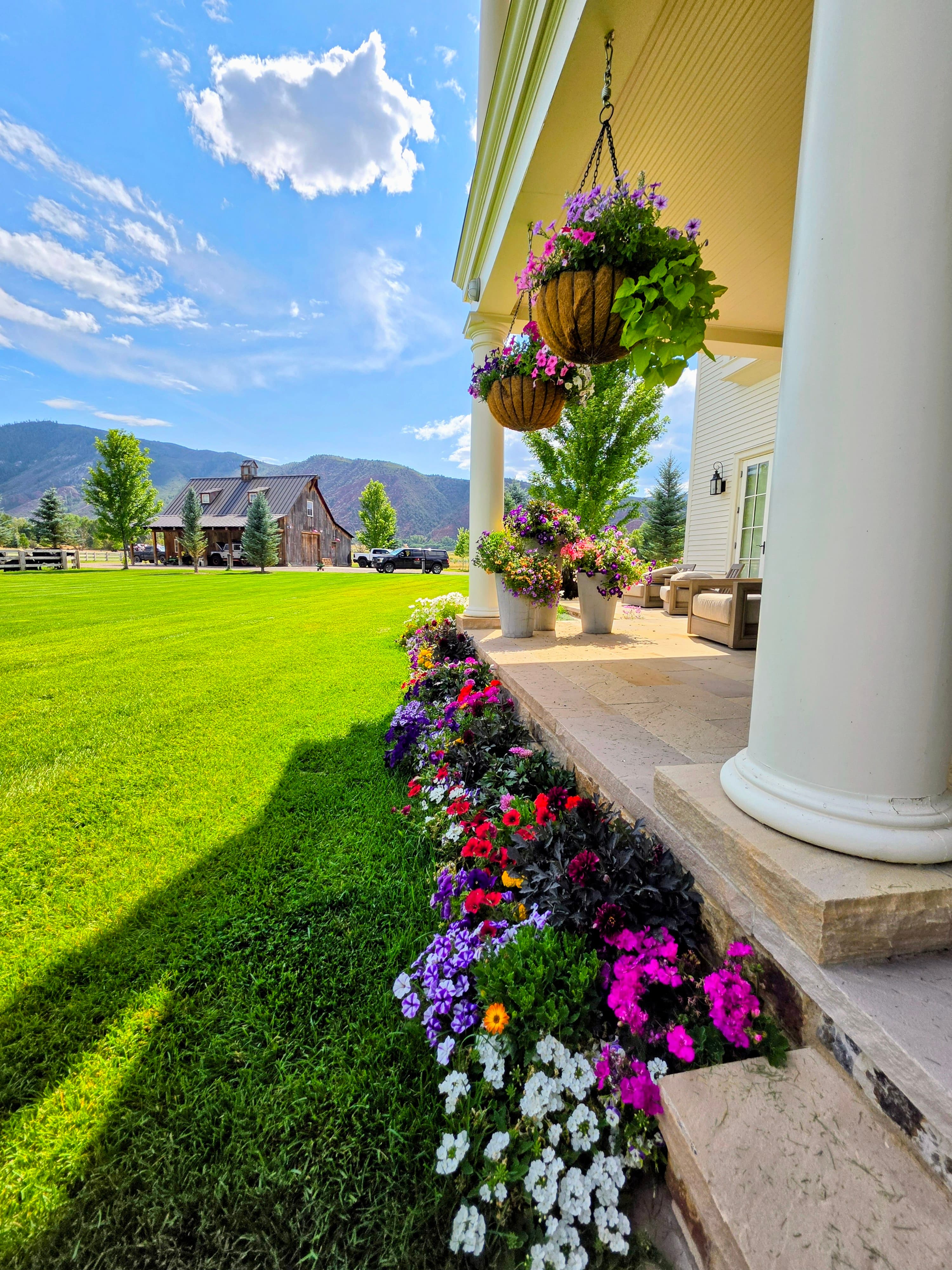 Colorful flowers line a white porch overlooking a green lawn, rustic barn, and distant mountains.